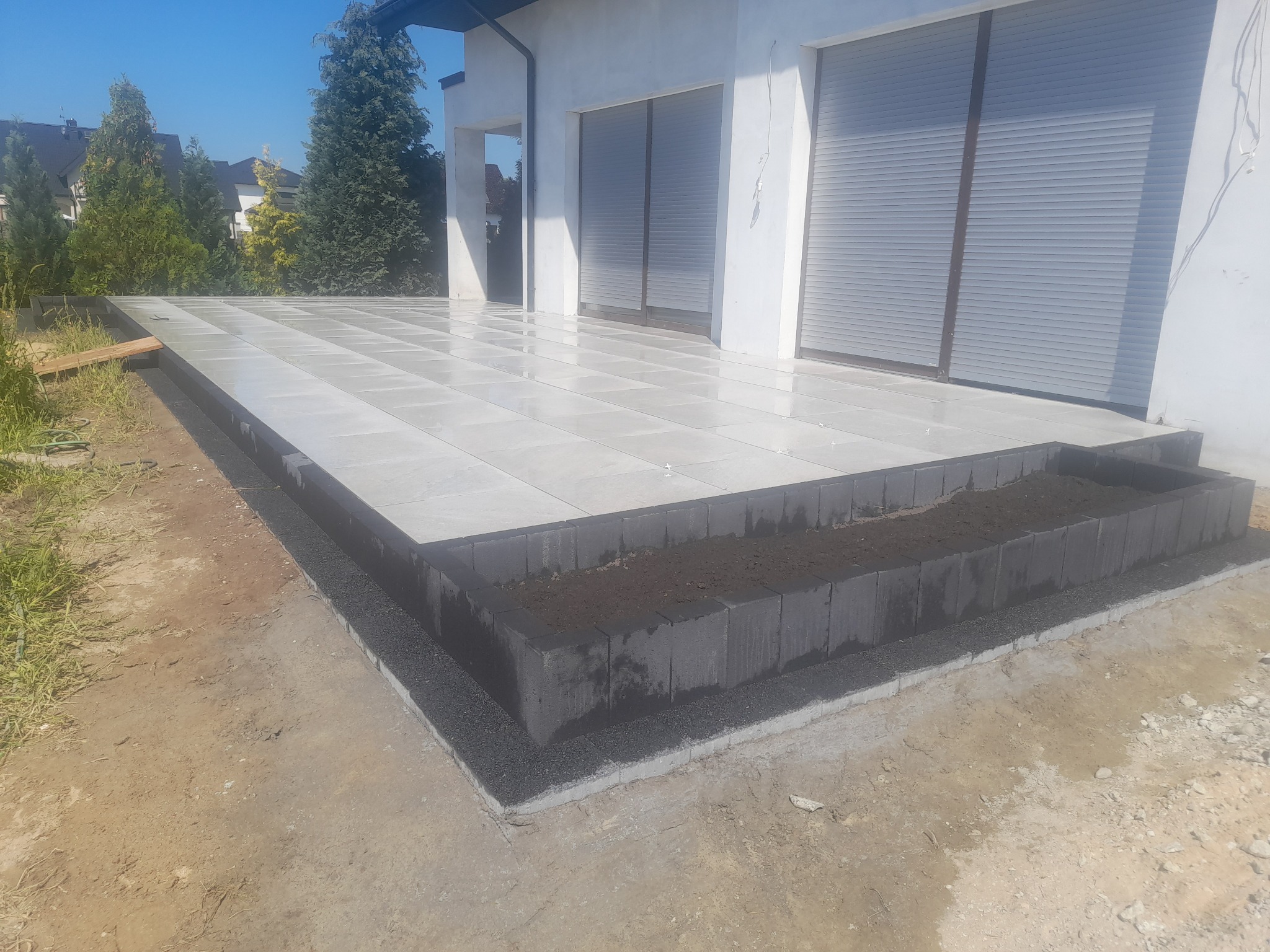 Jasna, nowa nawierzchnia tarasu z płyt betonowych, obrzeże z kostki w kolorze grafitowym, przygotowane pod nasadzenia roślin; widok na elewację domu z roletami zewnętrznymi w tle, w słoneczny dzień.