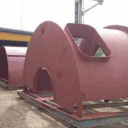 Czerwone, metalowe segmenty hali na wózkach transportowych, z widocznymi otworami i elementami konstrukcyjnymi, czekają na montaż w hali produkcyjnej.