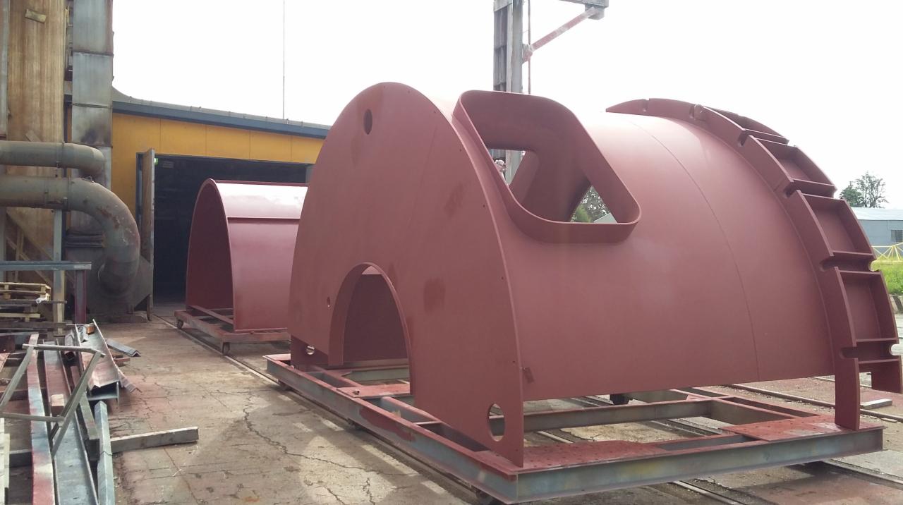 Czerwone, metalowe segmenty hali na wózkach transportowych, z widocznymi otworami i elementami konstrukcyjnymi, czekają na montaż w hali produkcyjnej.