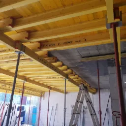 Żółte deskowanie stropu podparte metalowymi stemplami, widoczna drabina aluminiowa i bloczki z betonu komórkowego.