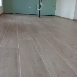 Ułożone płytki podłogowe imitujące drewno, jasne fugi, ściany częściowo wykończone płytami gipsowo-kartonowymi, widoczne instalacje.