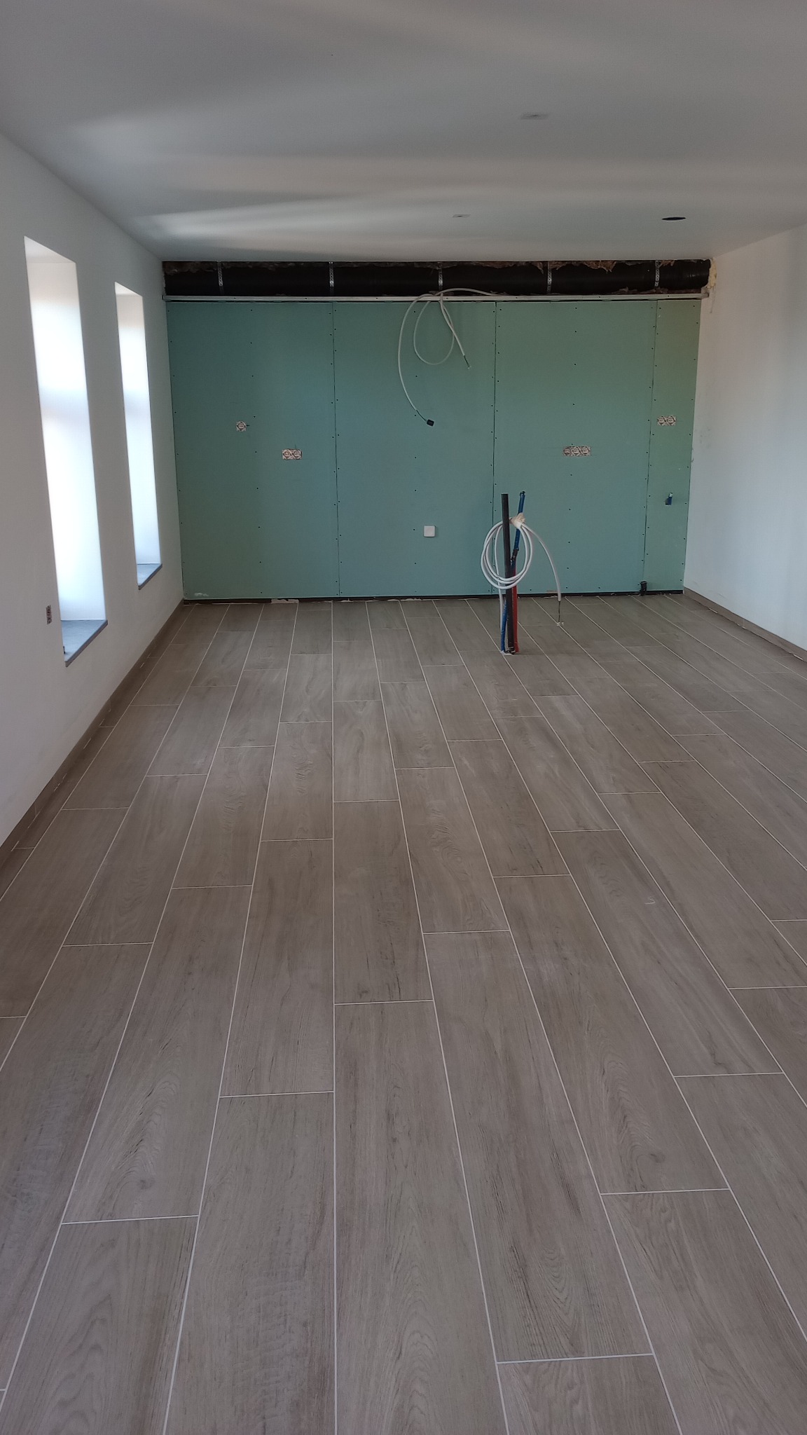 Wnętrze w trakcie prac wykończeniowych: położona podłoga z płytek imitujących drewno, ściana z płyt kartonowo-gipsowych z widocznymi przewodami instalacji wodnej i elektrycznej.