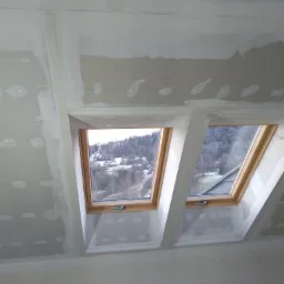 Dwa okna dachowe w trakcie montażu w poddaszu, widoczne płyty kartonowo-gipsowe i szpachlowane łączenia, zimowy krajobraz za oknami.