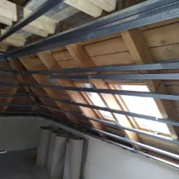 Stelaż z profili metalowych zamontowany do drewnianej konstrukcji poddasza, widoczne okno dachowe i rolki materiału izolacyjnego.