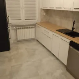 Wykończona kuchnia z białymi szafkami, drewnianym blatem, czarnym zlewem i szarymi płytkami podłogowymi, widok na okno z żaluzjami.