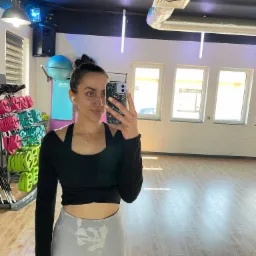 Kobieta robi selfie w sali ćwiczeń z hantlami i piłką gimnastyczną w tle, ubrana w sportowy strój.