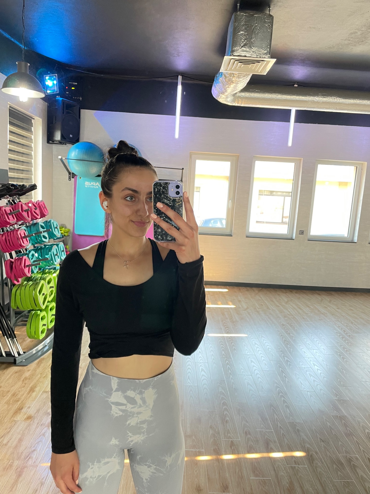 Kobieta robi selfie w sali ćwiczeń z hantlami i piłką gimnastyczną w tle, ubrana w sportowy strój.