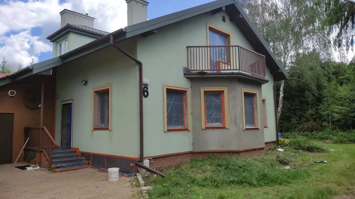 Dwupiętrowy dom z balkonem w trakcie prac elewacyjnych, okna zabezpieczone folią, widoczny numer domu '6' oraz częściowo nałożona warstwa szarej zaprawy.