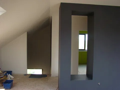Wnętrze w trakcie prac wykończeniowych: szara ściana z prostokątnym otworem, widok na pomieszczenie z oknem i zielonymi szafkami, fragment poddasza z oknem i narzędzia w wiadrach na podłodze.