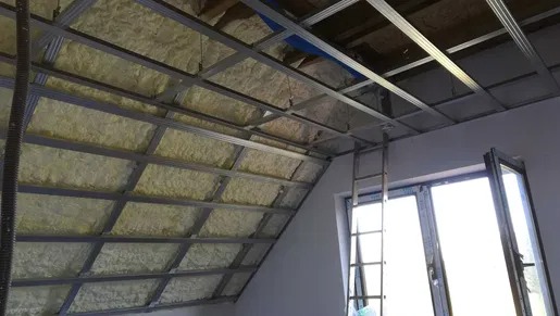 Montaż metalowego stelażu sufitowego z izolacją z wełny mineralnej na poddaszu, widoczna drabina oparta o ścianę przy oknie.