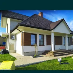 Nowoczesny dom jednorodzinny z brązowym dachem, elewacją w kolorze złamanej bieli i drewnianymi akcentami, widoczny fragment ocieplenia na ścianie bocznej, kostka brukowa na podejściu i trawnik.