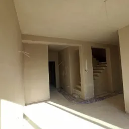 Wnętrze domu w stanie deweloperskim, ściany pokryte świeżo nałożoną, jasną warstwą, widoczne wystające kable elektryczne i schody w głębi.