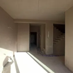 Wnętrze w trakcie prac wykończeniowych: świeżo otynkowane ściany w odcieniach beżu, widoczne kable elektryczne i schody w tle, silne światło słoneczne padające na podłogę.