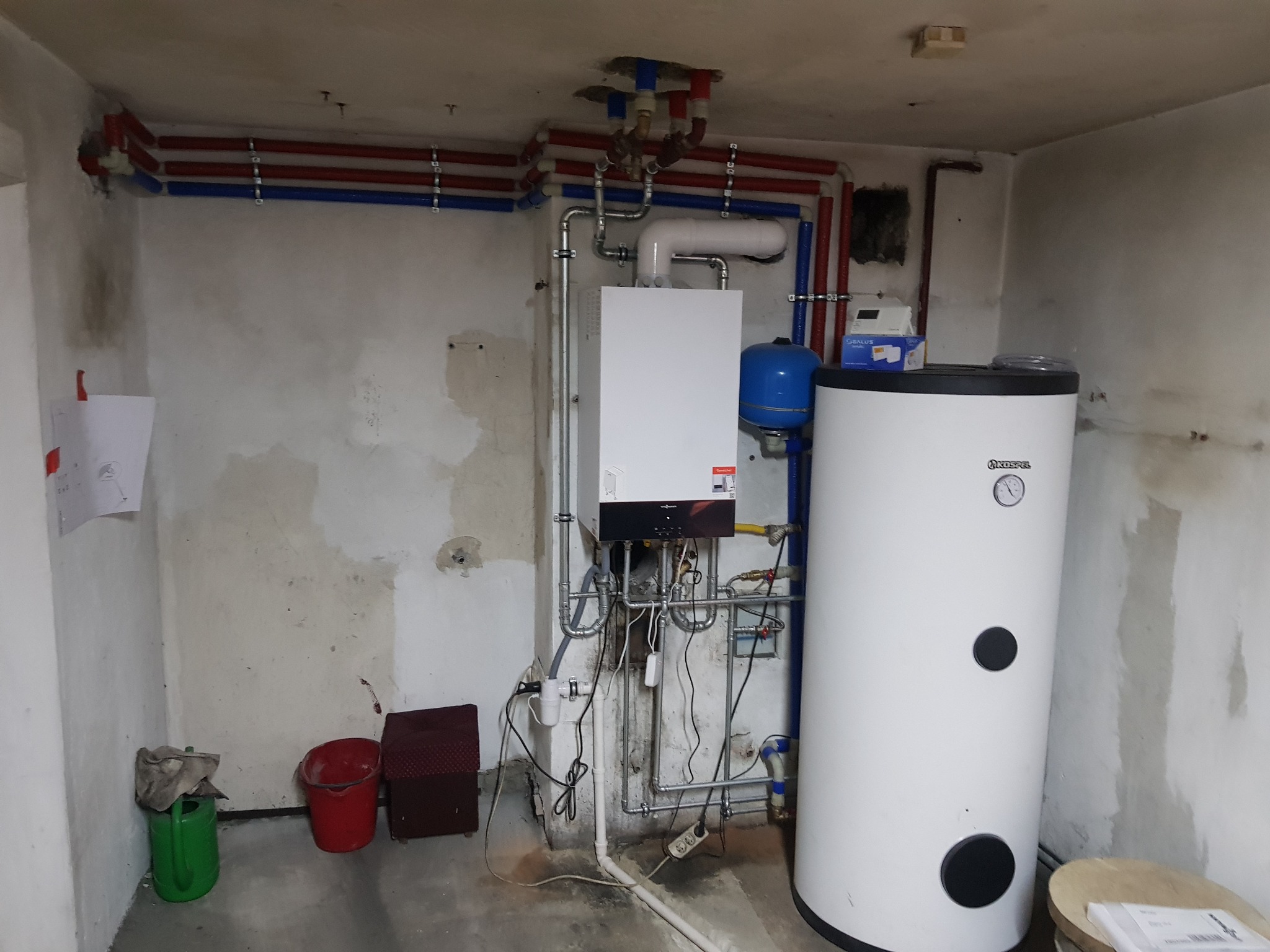 Kotłownia w trakcie prac instalacyjnych: biały piec gazowy Viessmann, zbiornik na wodę Kospel, instalacja z rur miedzianych i plastikowych w kolorach niebieskim i czerwonym, surowe ściany w tle...