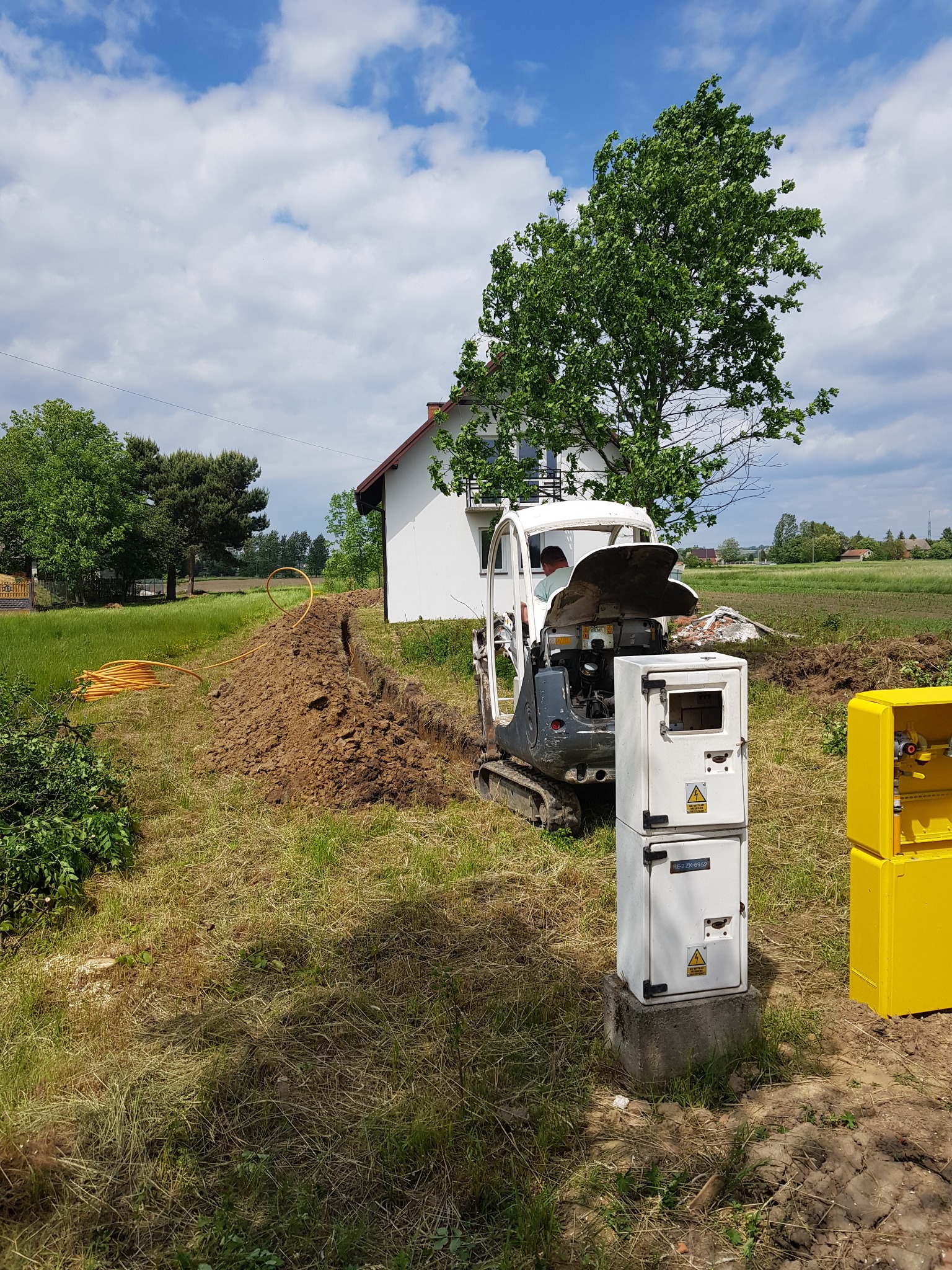 Mini koparka wykopuje rów pod żółty przewód gazowy na tle wiejskiego krajobrazu z białą skrzynką gazową i żółtą szafką gazową.