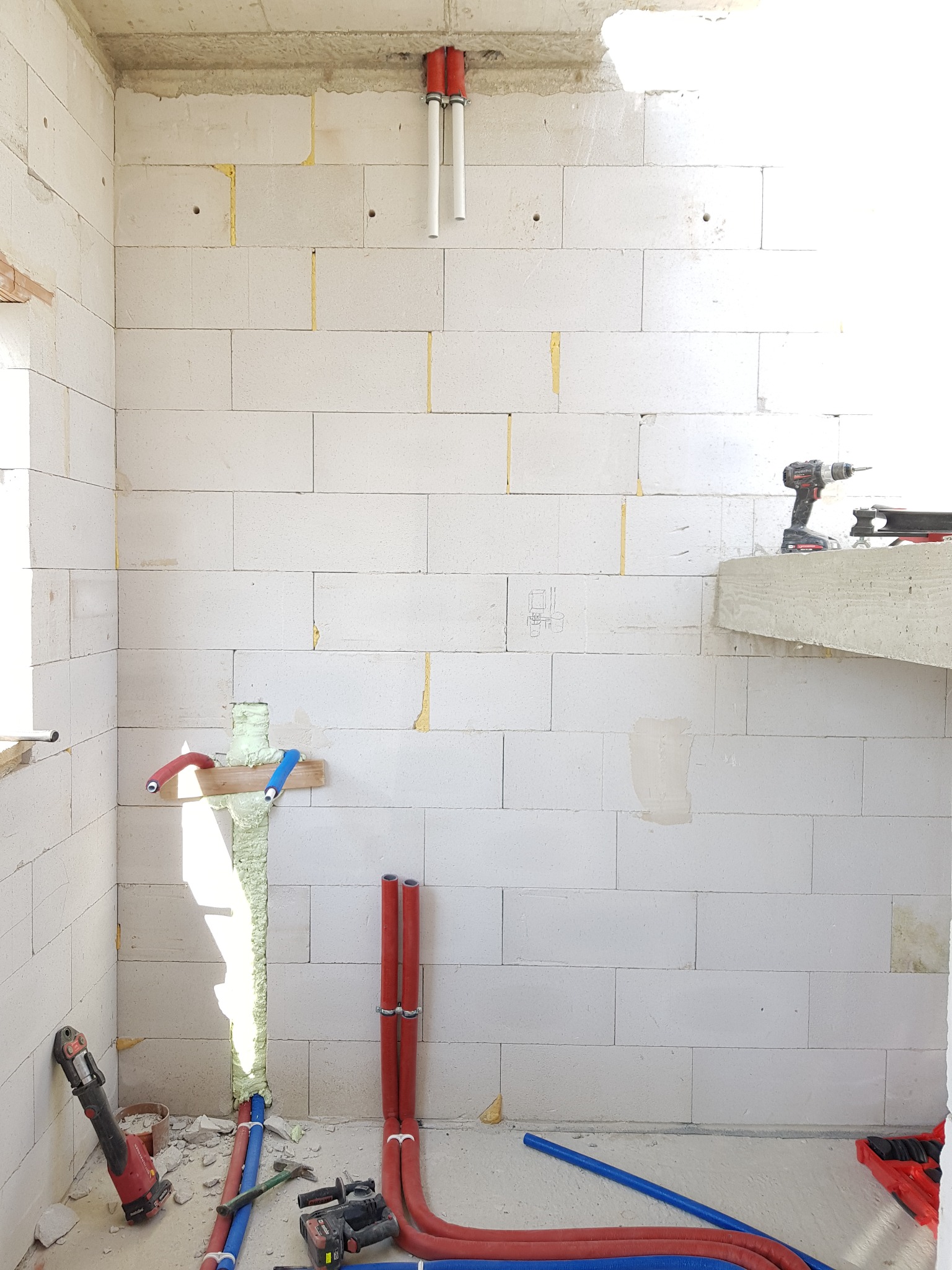 Surowe wnętrze w trakcie instalacji hydraulicznej: widoczne białe ściany z betonu komórkowego, pionowe rury w ścianie, narzędzia hydrauliczne na podłodze i szczelina wypełniona pianką montażową.
