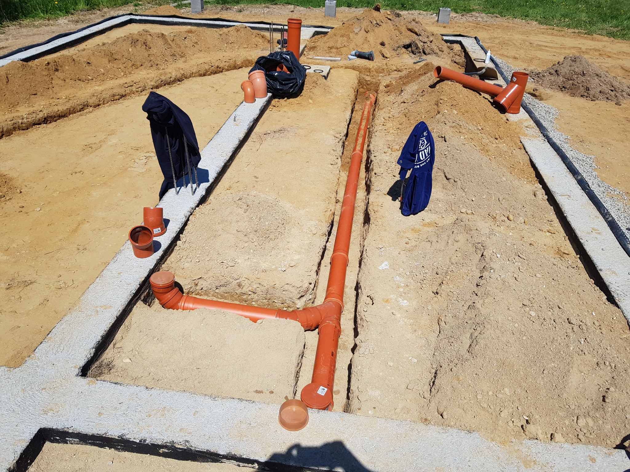 Fundamenty domu z wykopanymi rowami pod instalacje sanitarne, widoczne pomarańczowe rury kanalizacyjne łączone kształtkami.