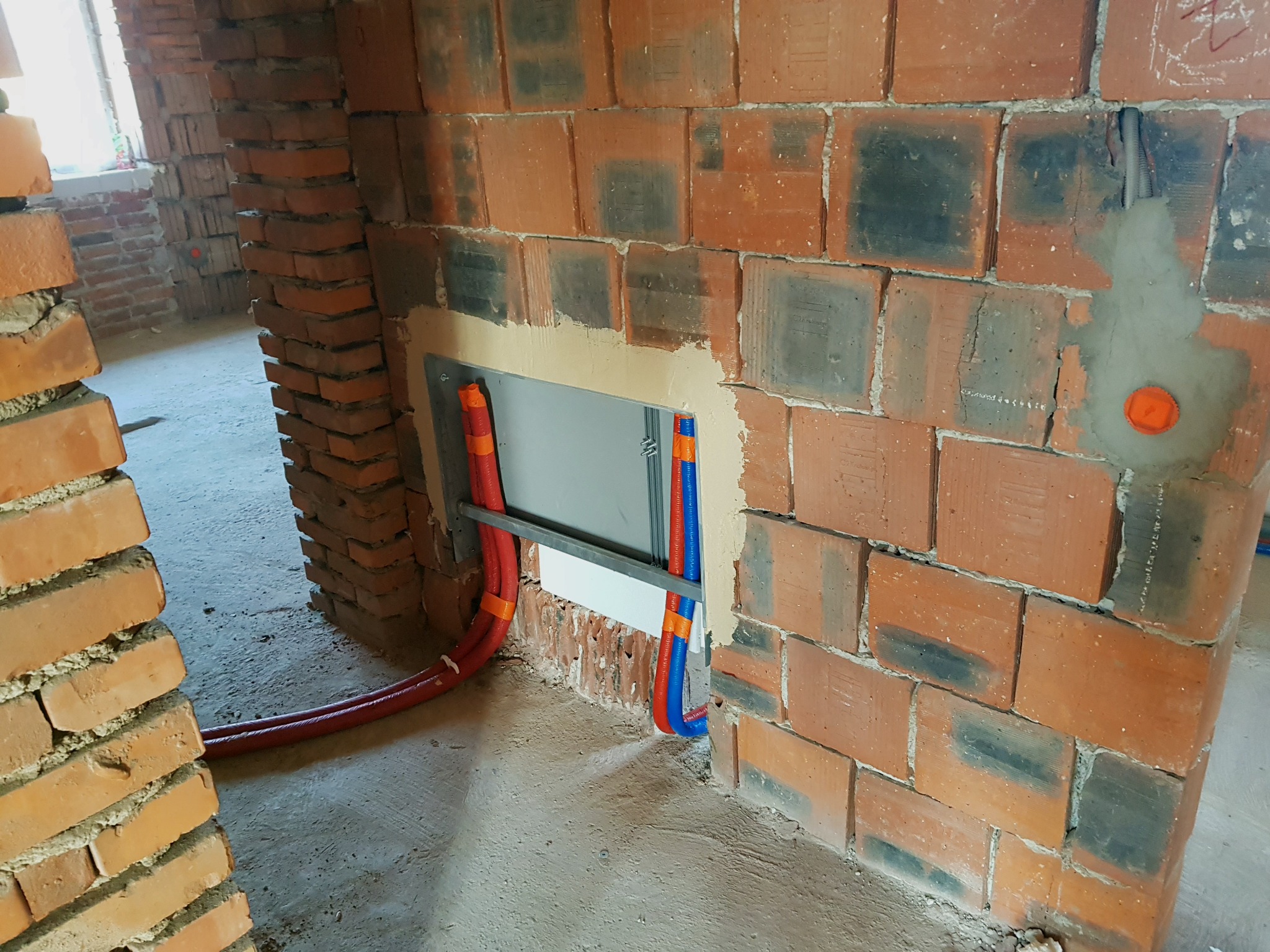 Fragment ściany z czerwonej cegły z widocznymi rurami instalacji wodnej w kolorze czerwonym i niebieskim, zabezpieczonymi pomarańczową taśmą, wychodzącymi z otworu w ścianie. Otwór wokół rur jest...
