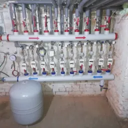 Rozdzielacz ogrzewania podłogowego z licznikami ciepła, zamontowany na ścianie z cegły, widoczne rury doprowadzające i odprowadzające wodę, szare izolacje na rurach, szary zbiornik wyrównawczy.