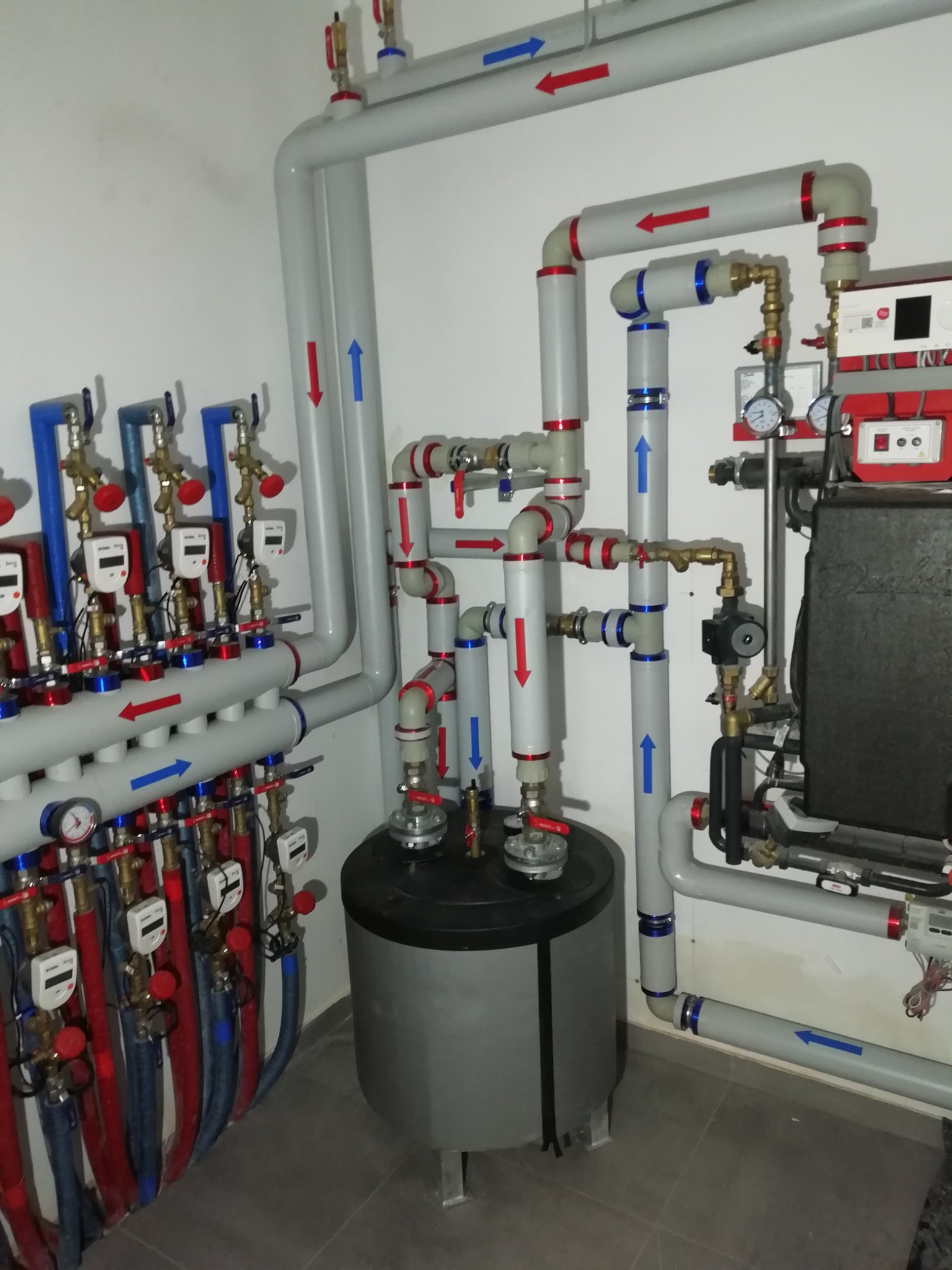 Rozbudowana instalacja grzewcza z rurami oznaczonymi strzałkami, kolektor z licznikami ciepła i zasobnik wody użytkowej w kotłowni.
