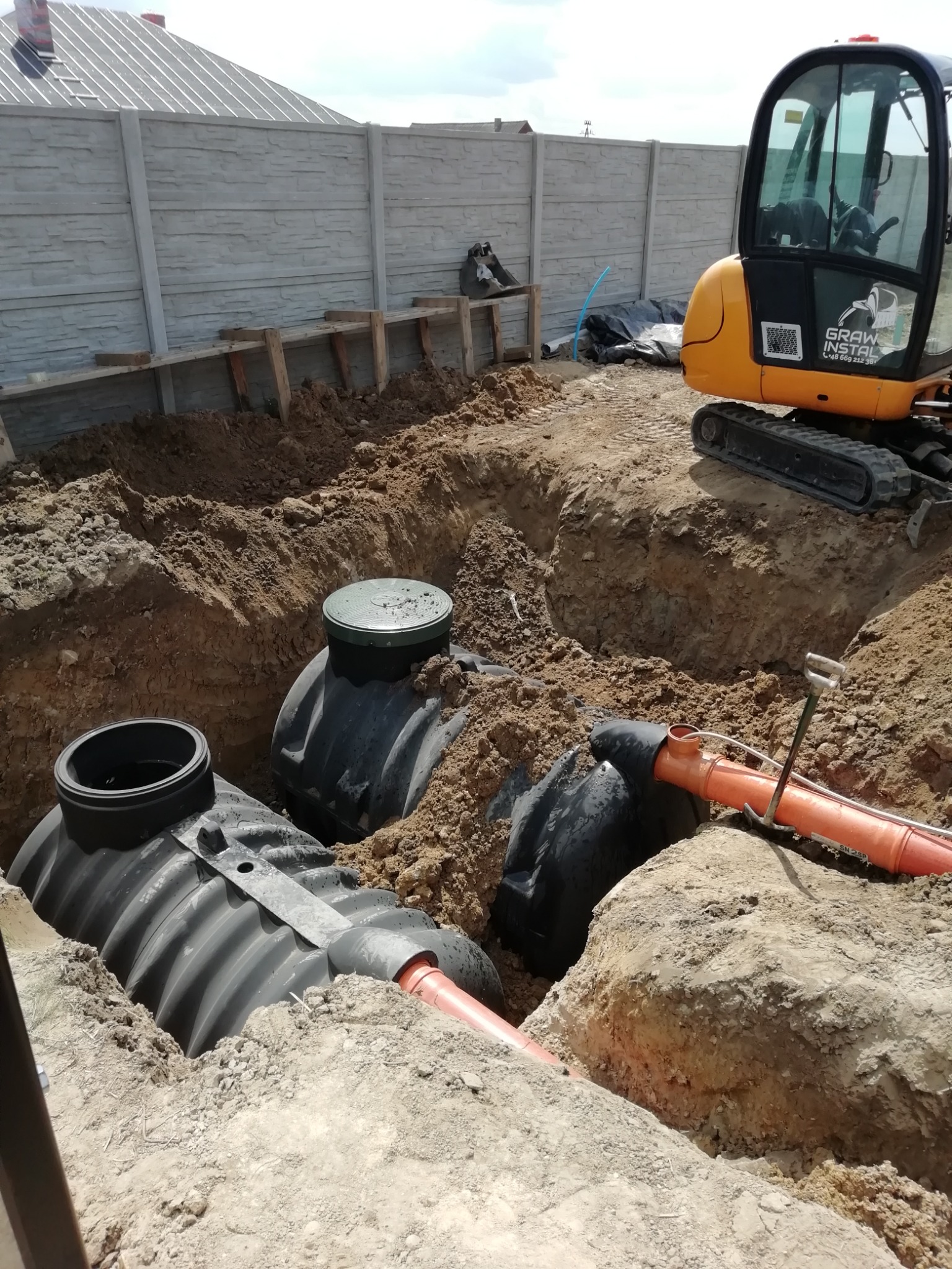 Montaż szamba ekologicznego z rurami kanalizacyjnymi w wykopie, widoczna mini koparka i fragment betonowego ogrodzenia.