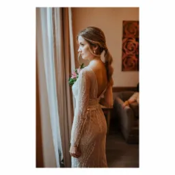 Panna młoda w koronkowej sukni z odkrytymi plecami, z włosami upiętymi w niski kucyk, spogląda przez okno trzymając bukiet kwiatów.