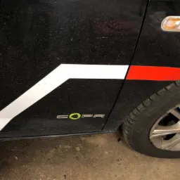 Boczny widok brudnego, czarnego samochodu z białymi i czerwonymi paskami oraz napisem 'COPA' z zielonym akcentem, na tle betonowej podłogi.