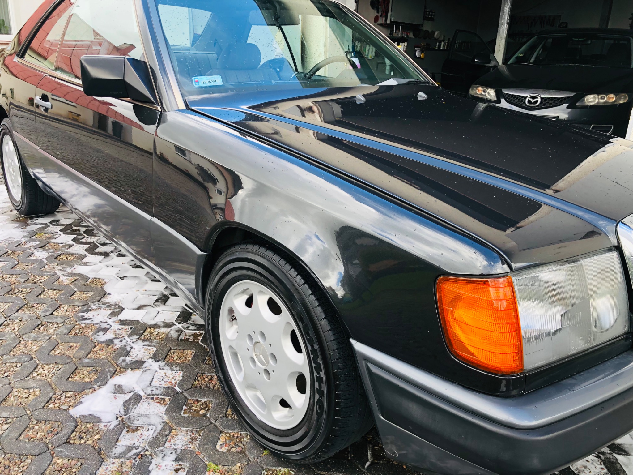 Czarny Mercedes-Benz coupe po umyciu, widoczny na kostce brukowej z resztkami piany, z zaparkowanym czarnym samochodem w tle.