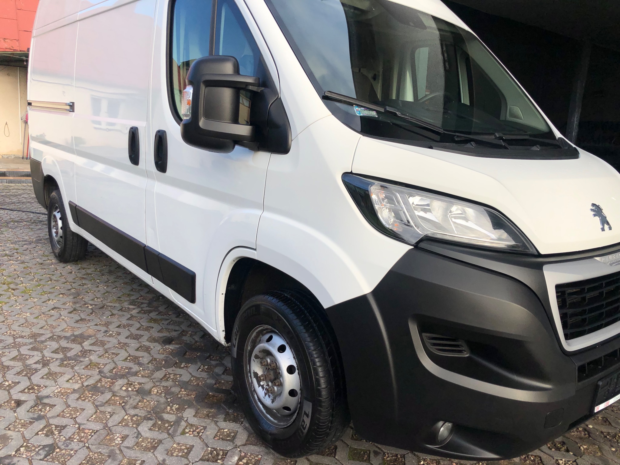 Biały samochód dostawczy Peugeot Boxer zaparkowany na brukowanym placu, widok z przodu i z boku, z widocznym logo marki na masce.