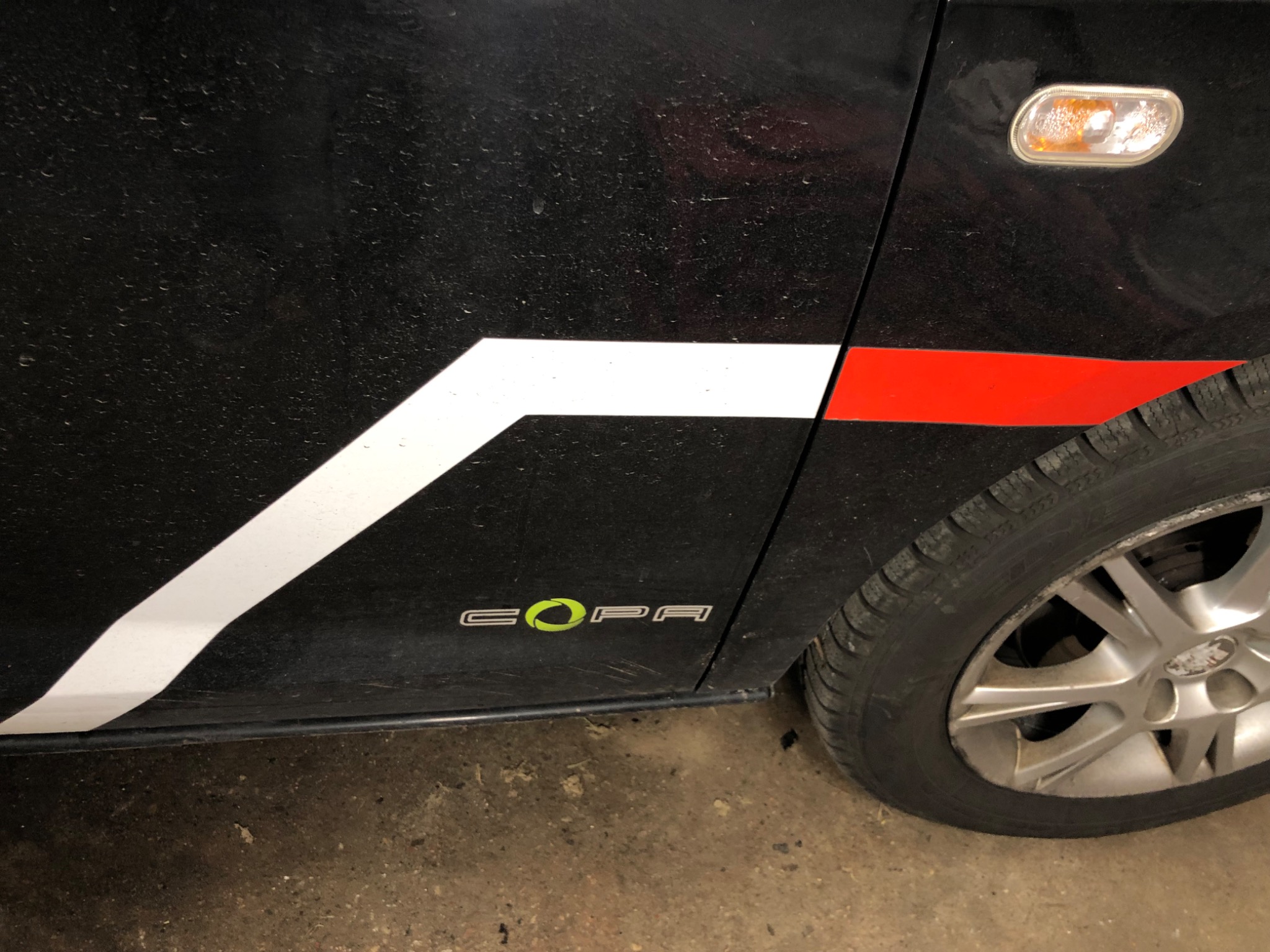 Boczny widok brudnego, czarnego samochodu z białymi i czerwonymi paskami oraz napisem 'COPA' z zielonym akcentem, na tle betonowej podłogi.