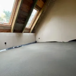 Wnętrze poddasza z wylaną, świeżą wylewką betonową, zabezpieczoną folią przy ścianach, widoczne okna dachowe z drewnianą ramą i częściowo wykończone ściany.