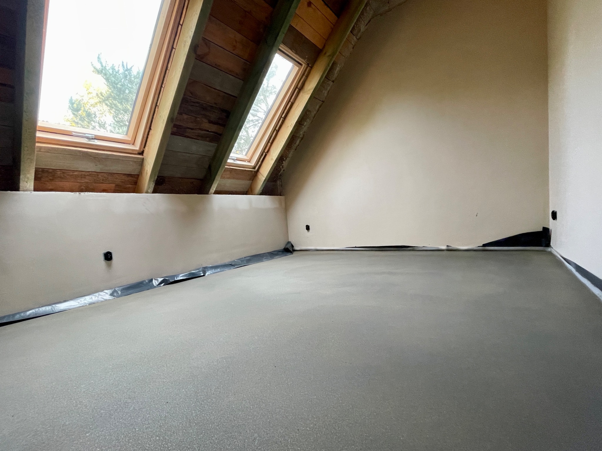 Wylewka betonowa na poddaszu z oknami dachowymi, zabezpieczona folią przy ścianach, gotowa do dalszych prac wykończeniowych.