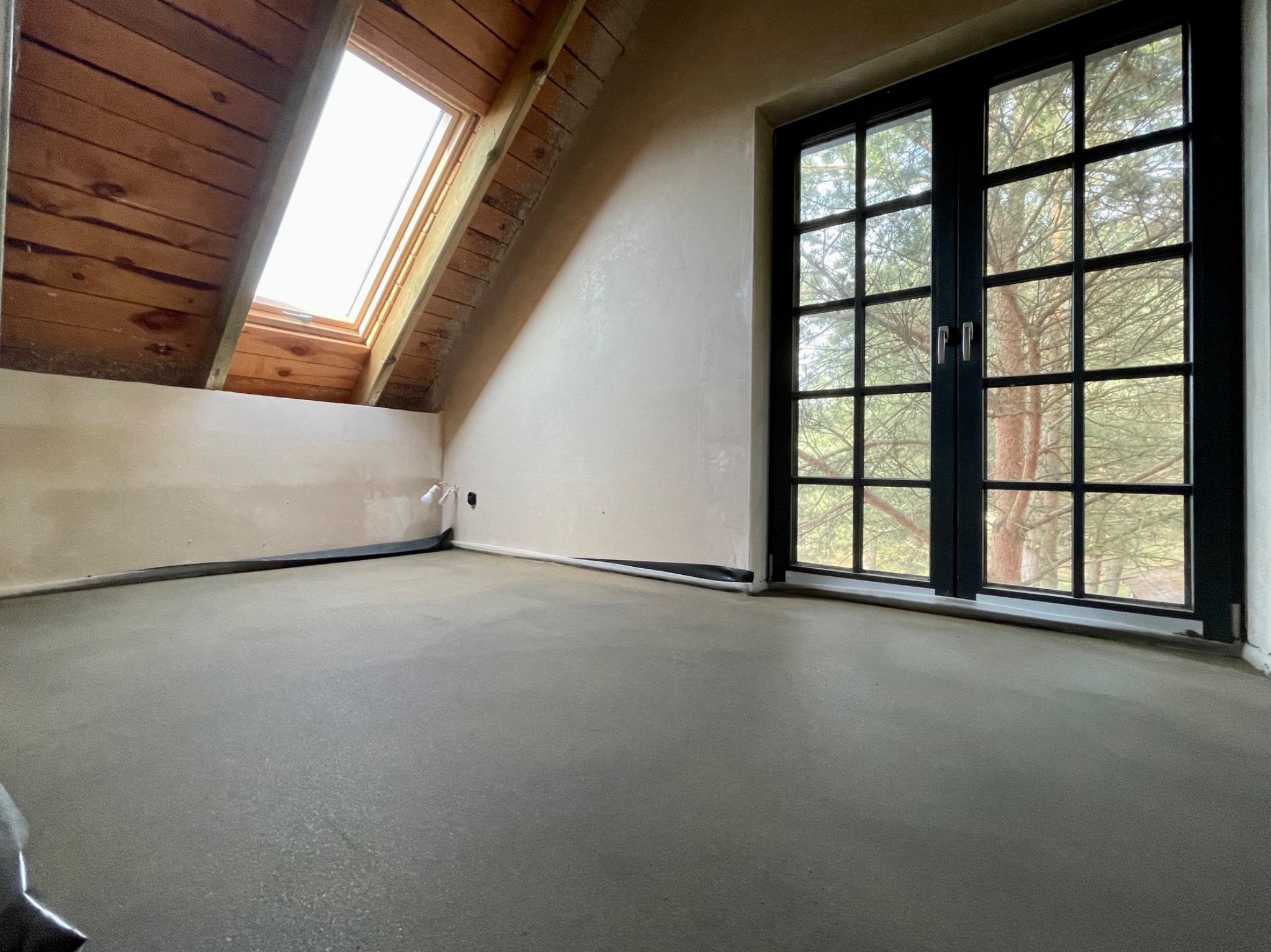 Poddasze z oknem dachowym i dużym oknem z czarną ramą, ściany po tynkowaniu, wylewka podłogowa w szarym kolorze.