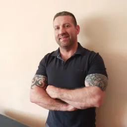 Portret mężczyzny z tatuażami na ramionach, w polo, skrzyżowane ręce, oparty o stół do zabiegów w Grodzisku Wielkopolskim.