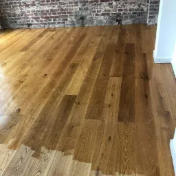 Częściowo odnowiona drewniana podłoga z widoczną różnicą w kolorze i wykończeniu, ceglana ściana w tle.