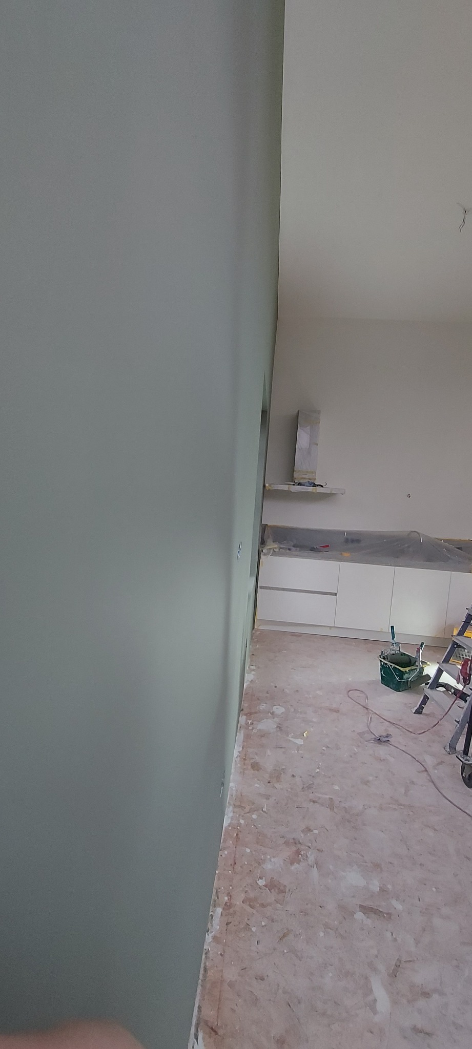 Świeżo pomalowana ściana w odcieniach szarości w pomieszczeniu w trakcie remontu, widoczne zabezpieczone meble kuchenne, drabina malarska i wiadro z farbą.
