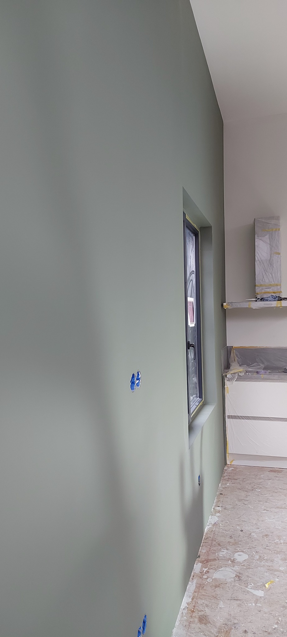 Świeżo pomalowana ściana w odcieniu zieleni z widocznymi zabezpieczonymi gniazdkami elektrycznymi i oknem z czarną ramą, fragment zabezpieczonej folią kuchni w tle.