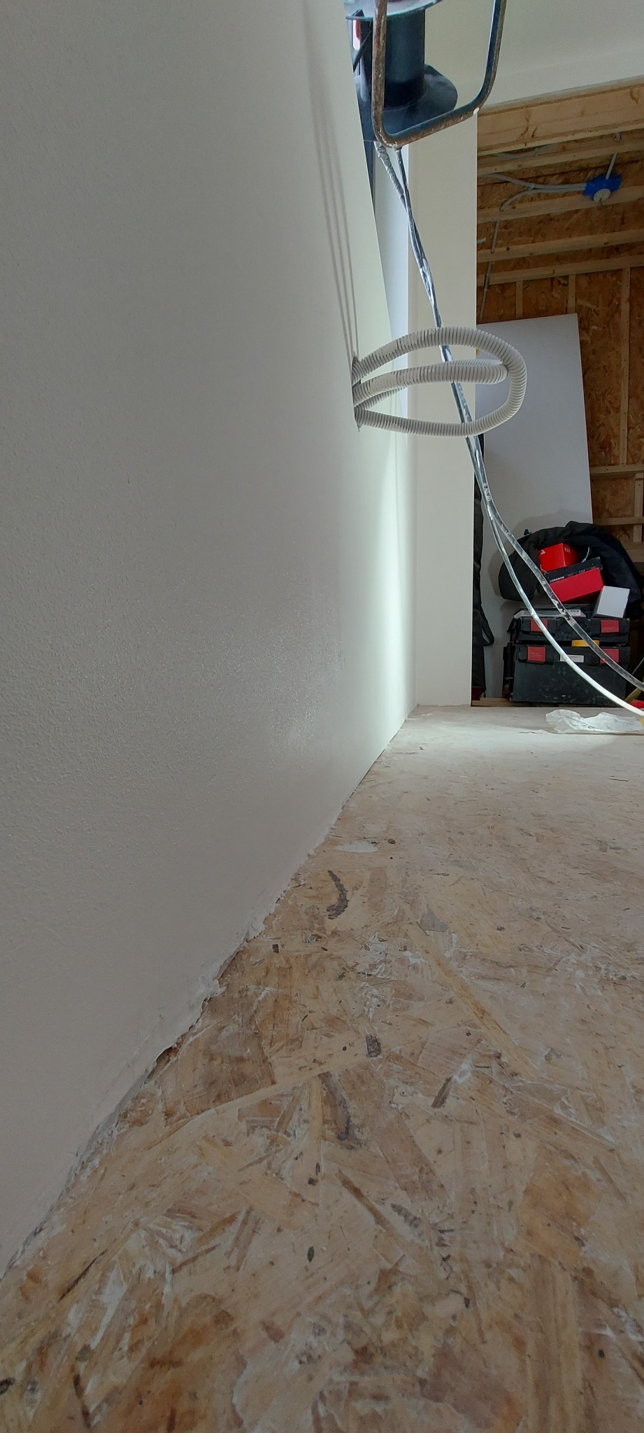 Świeżo pomalowana ściana w trakcie remontu, widoczne kable w peszlu wychodzące ze ściany, surowa podłoga z płyty OSB, narzędzia w tle.