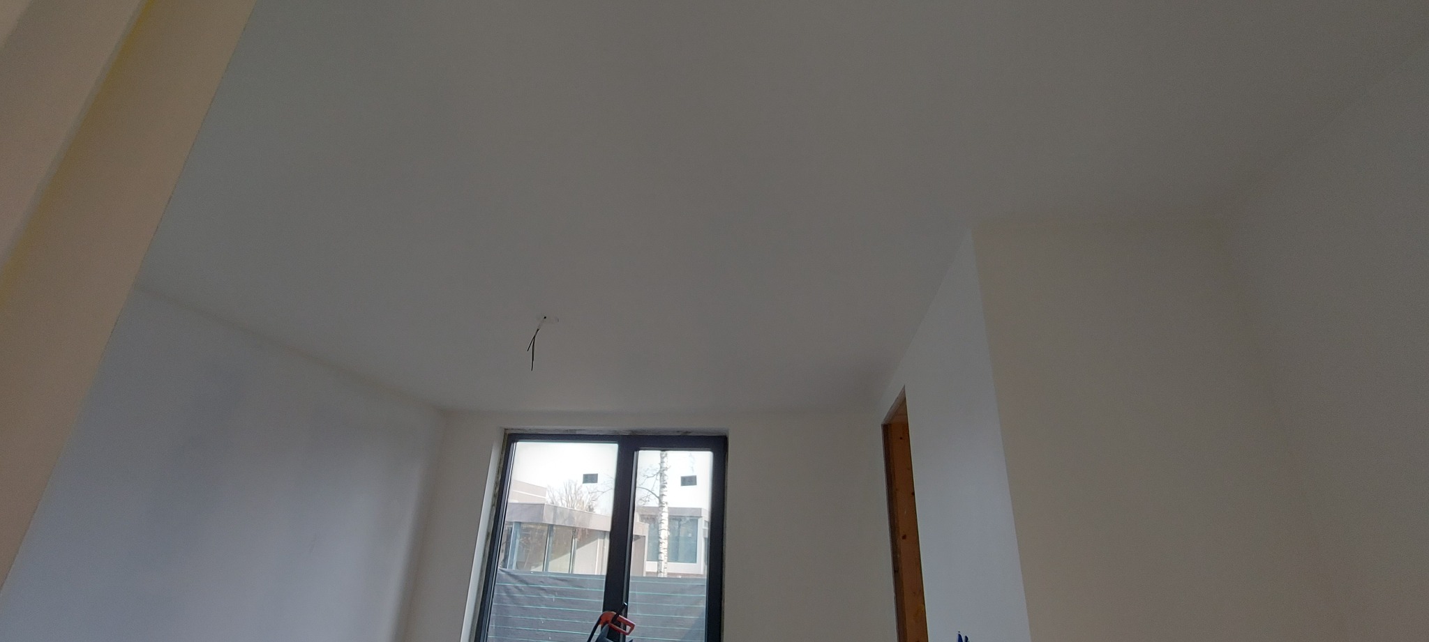 Wnętrze świeżo wykończonego pokoju z białymi ścianami i sufitem, widoczne okno balkonowe z widokiem na zewnątrz, wystające kable ze ściany, przygotowane do montażu oświetlenia.