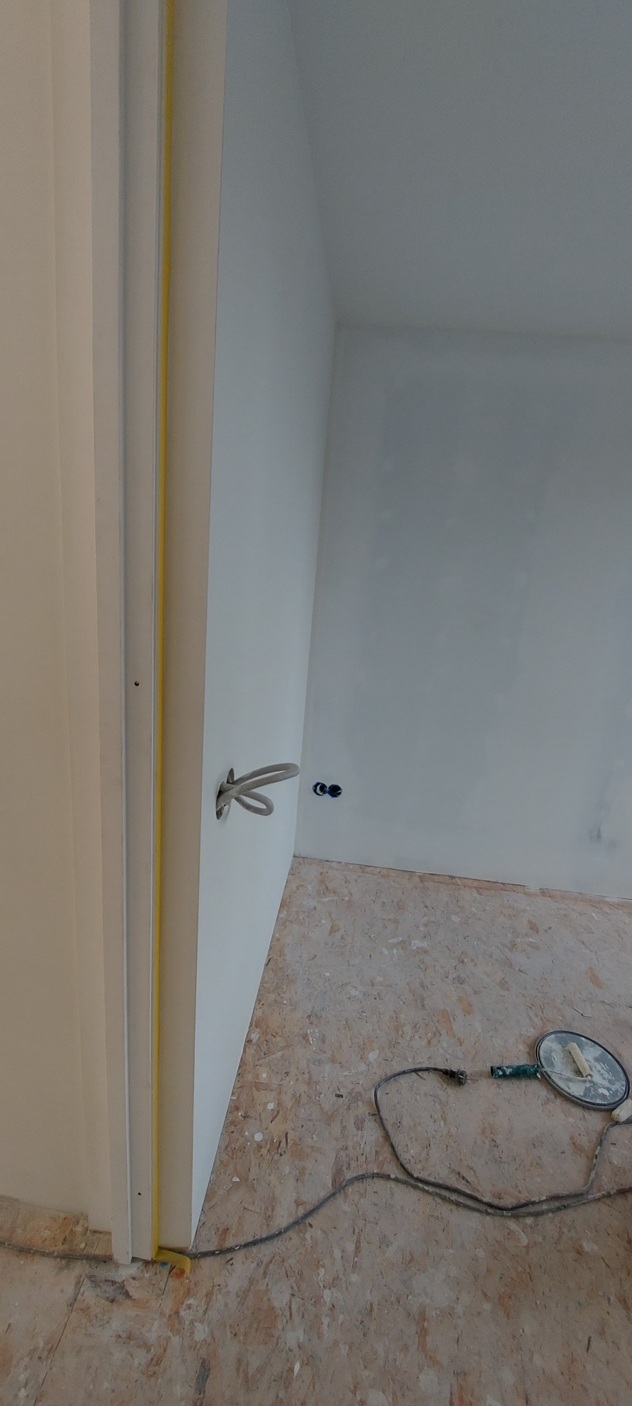Świeżo wyszpachlowana, biała ściana z widocznymi gniazdkami elektrycznymi i wystającymi kablami, zabezpieczona żółtą taśmą malarską przy framudze drzwi, na podłodze leży szlifierka do gładzi.