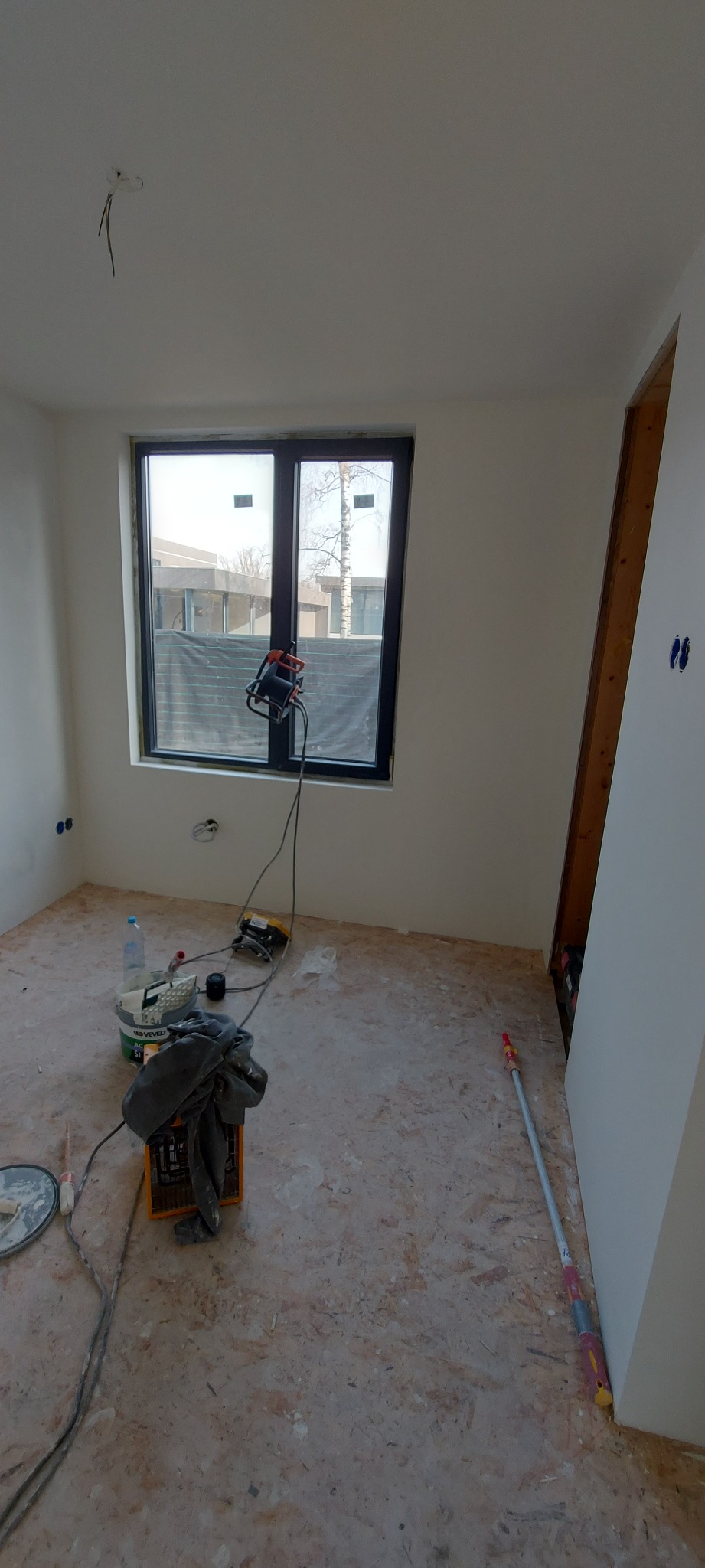 Pokój w trakcie remontu, ściany pomalowane na biało, widoczne narzędzia malarskie, szlifierka do gładzi oparta o okno, na podłodze wiadro z farbą i wałek do malowania.