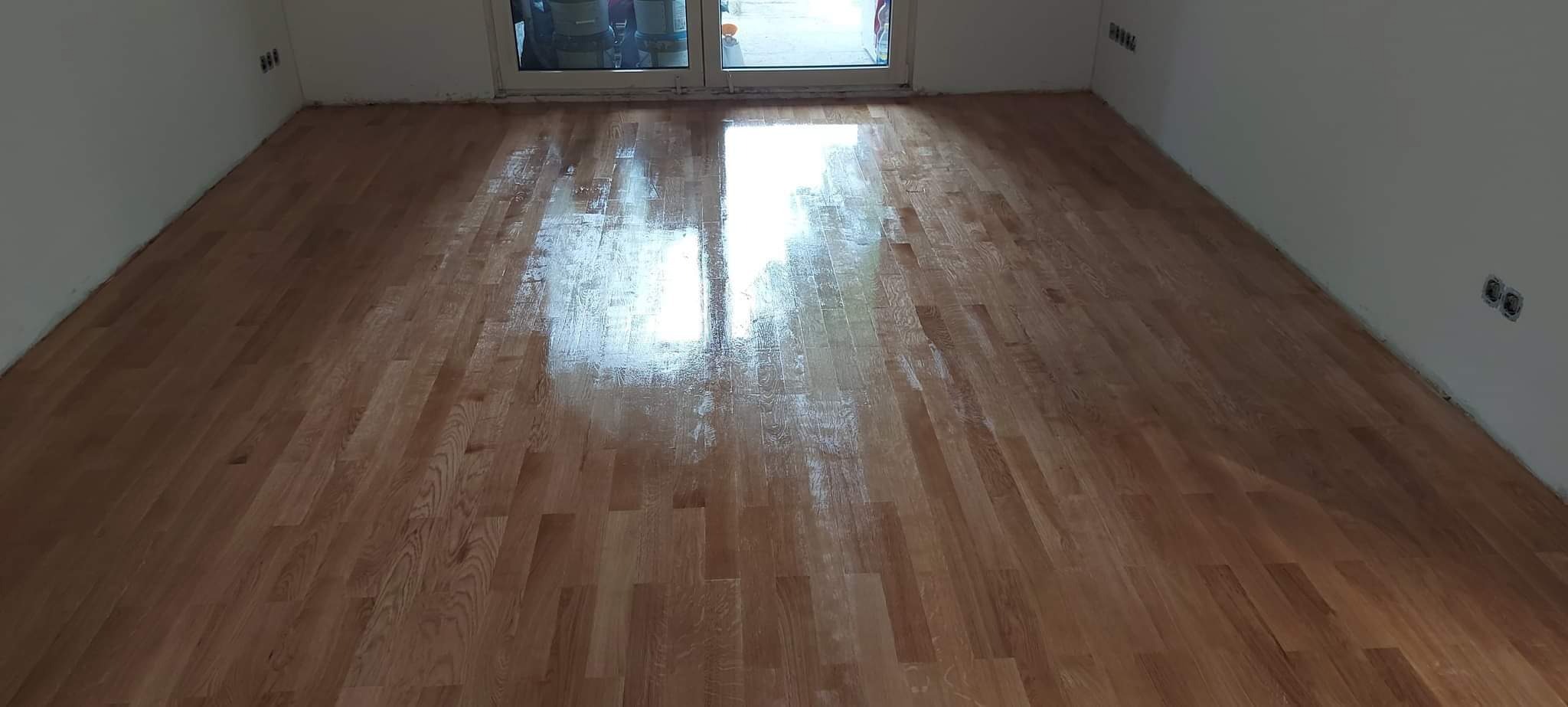 Świeżo położony, lakierowany parkiet dębowy w pustym pokoju z białymi ścianami, widoczne gniazdka elektryczne i okno balkonowe z pojemnikami farb w tle.