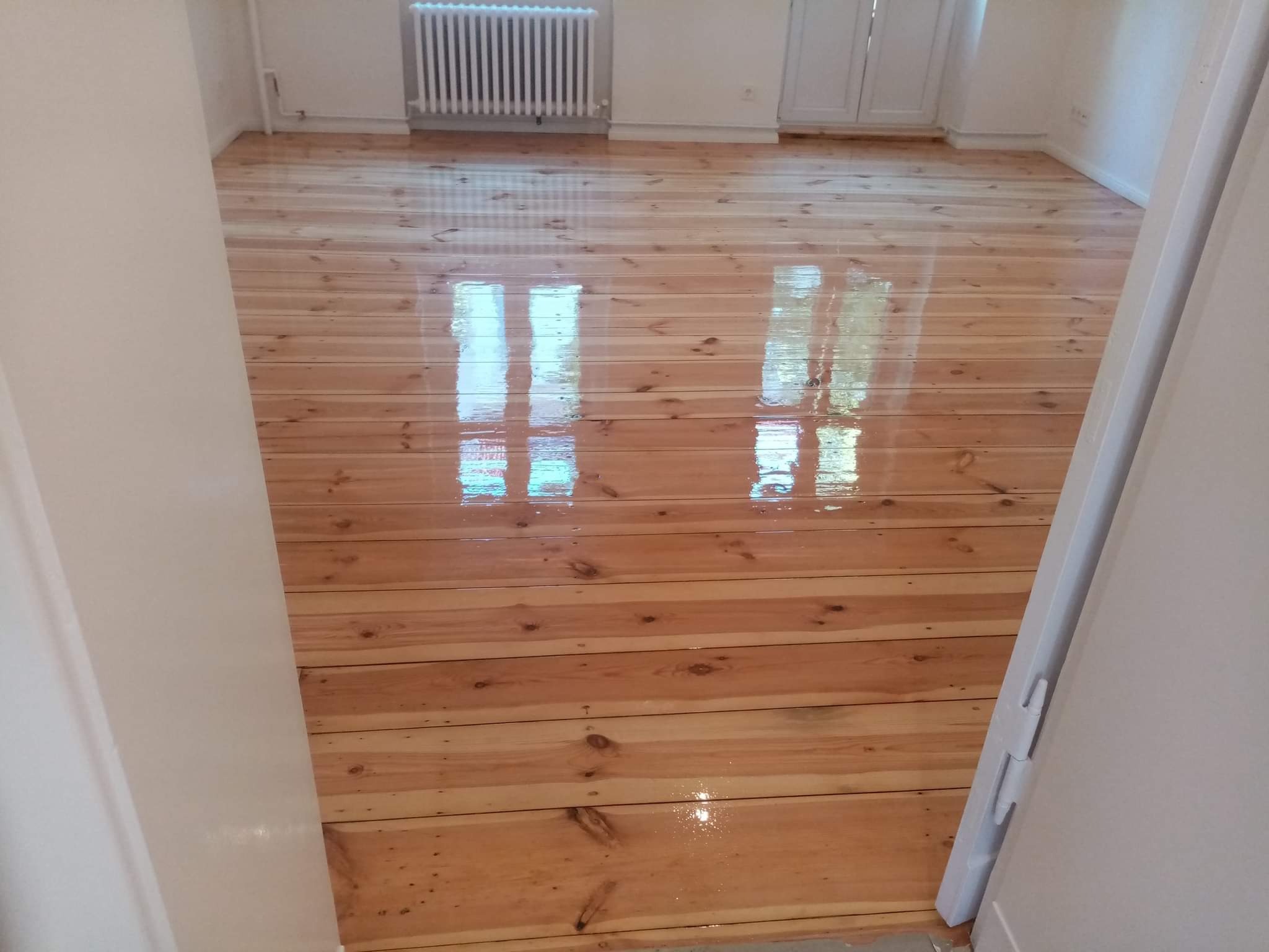 Świeżo polakierowana, błyszcząca podłoga z desek w jasnym odcieniu drewna, widoczna zza otwartych drzwi w jasnym wnętrzu z białym grzejnikiem.