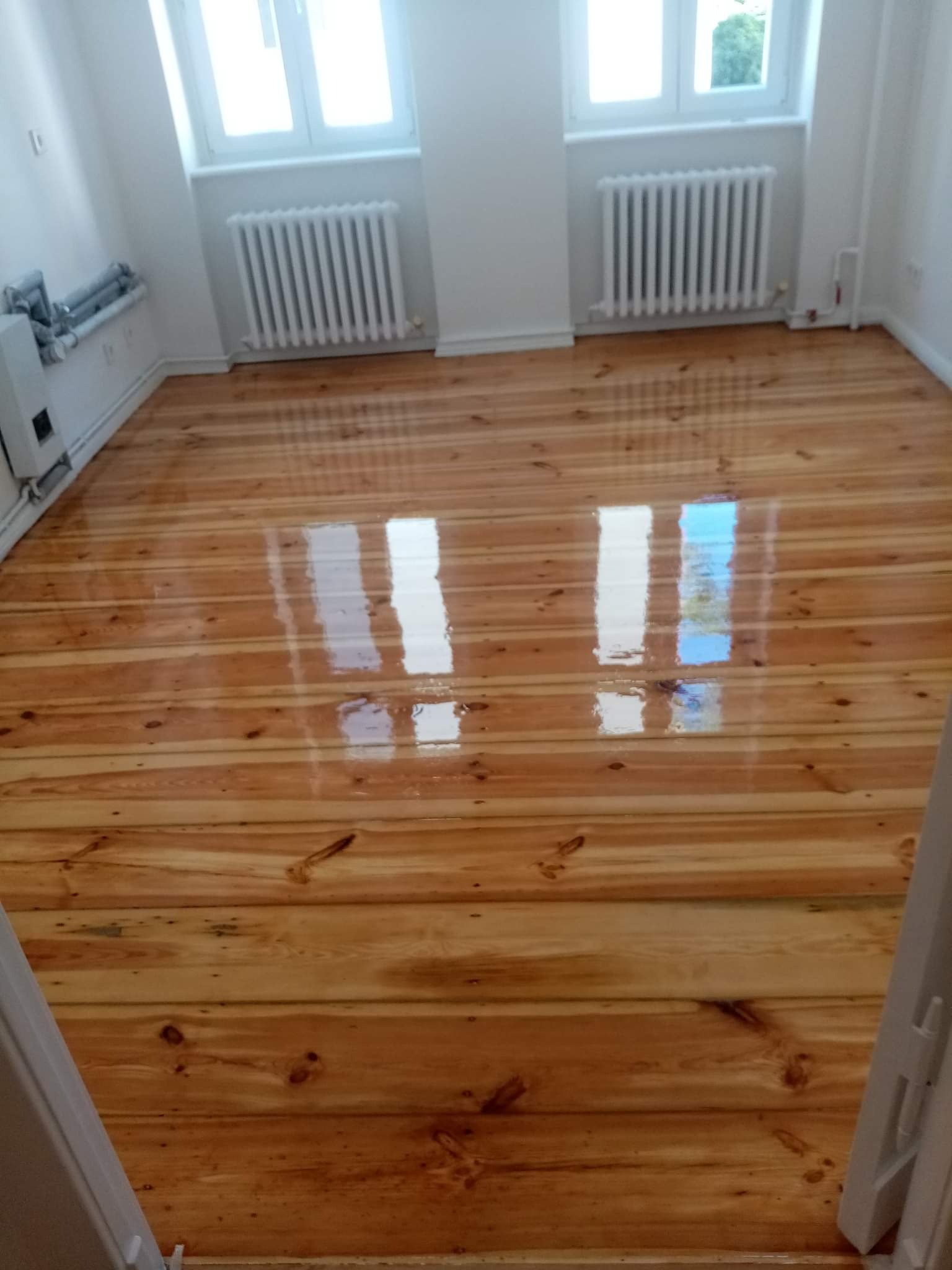 Błyszcząca, świeżo polakierowana podłoga z desek w jasnym pomieszczeniu z białymi ścianami i grzejnikami, widoczne odbicia okien.