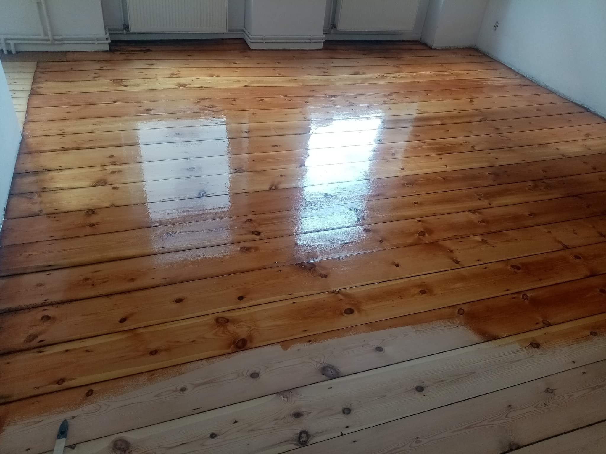 Fragment drewnianej podłogi w trakcie lakierowania; połowa powierzchni pokryta lakierem o ciepłym, brązowym odcieniu, druga połowa w surowym, jasnym drewnie, widoczny pędzel w rogu.