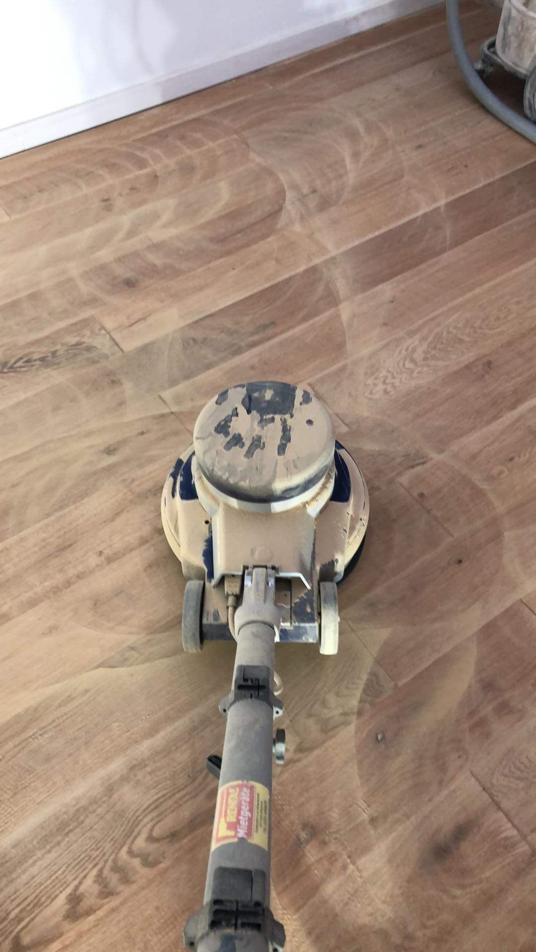 Podczas renowacji parkietu w Gnieźnie, widoczna maszyna do obróbki drewna, pozostawiająca charakterystyczne ślady po cyklinowaniu, z widocznym pyłem i resztkami lakieru na powierzchni.