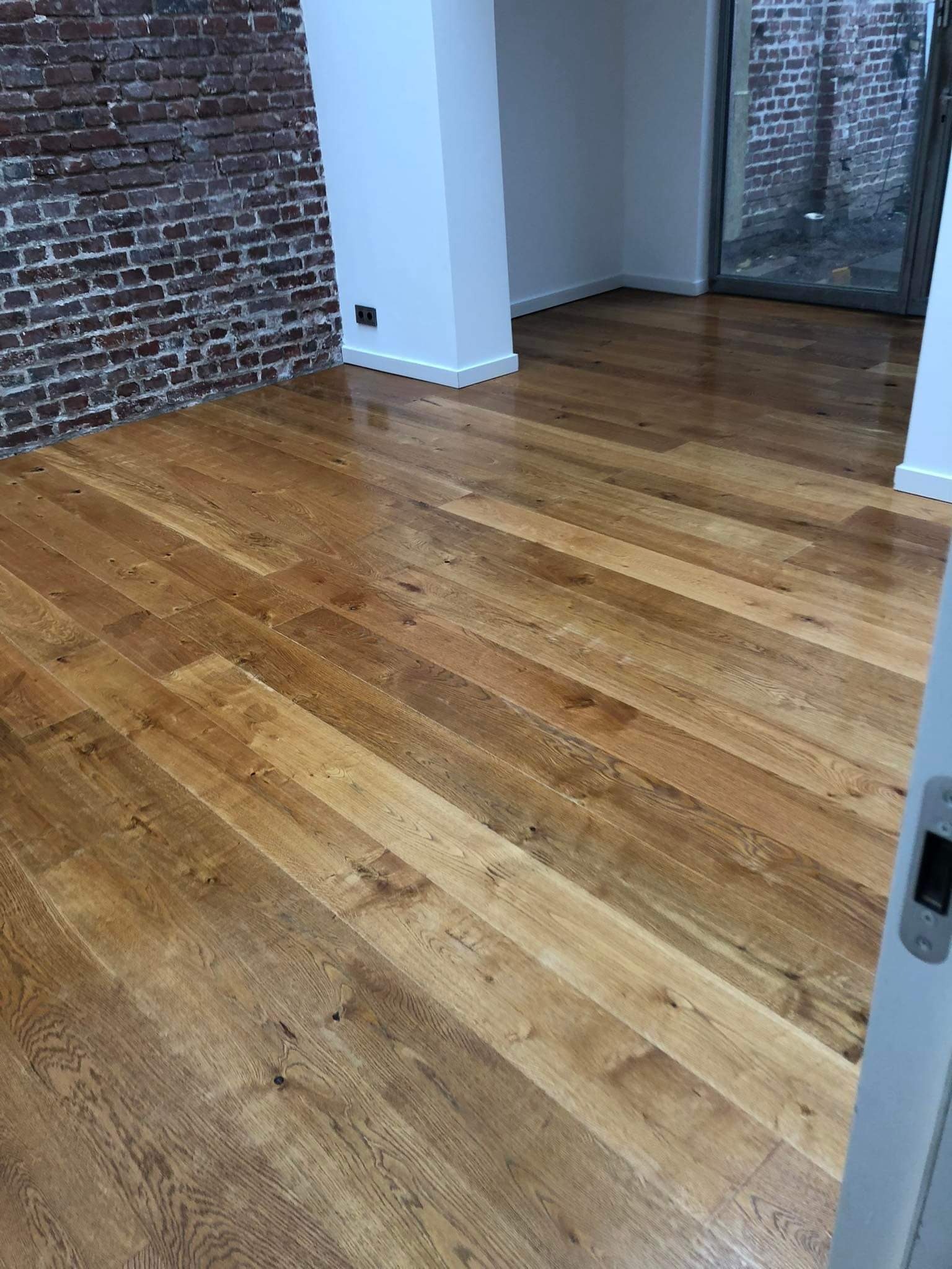 Nowo ułożony, lakierowany parkiet z naturalnego drewna w pomieszczeniu z ceglaną ścianą i białymi listwami przypodłogowymi.