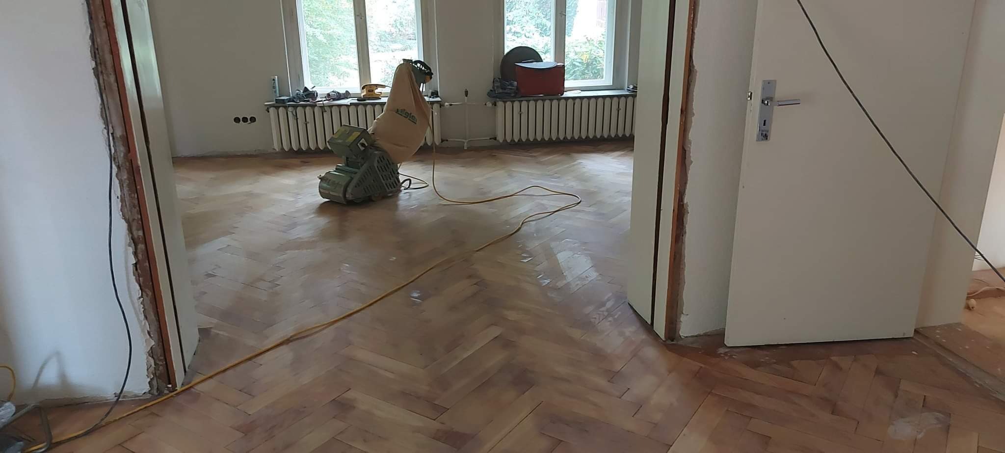 Parkiet w jodełkę częściowo odnowiony, z widoczną maszyną do szlifowania i workiem na pył, wnętrze w trakcie remontu z odsłoniętymi framugami drzwi.