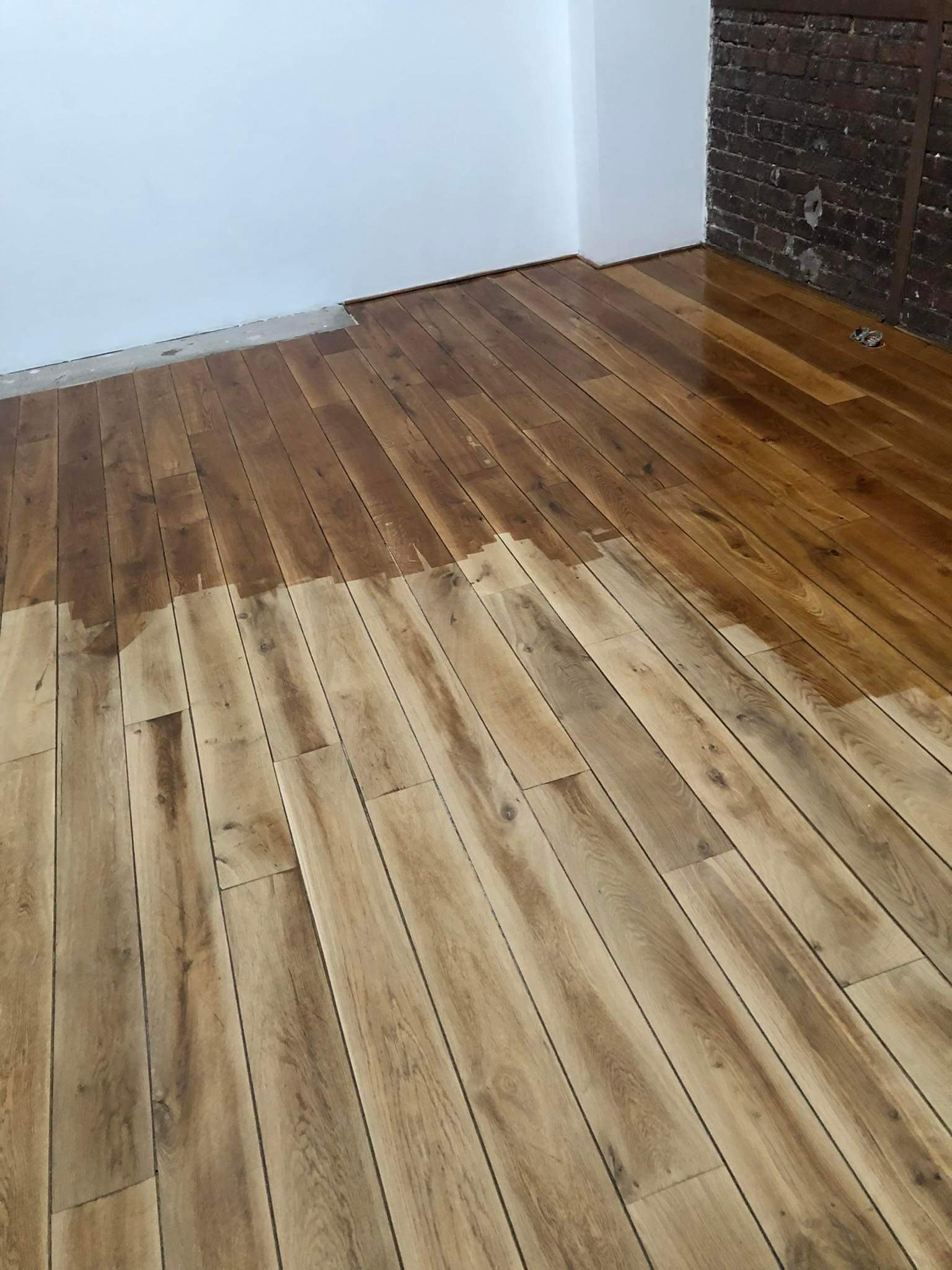 Częściowo polakierowany parkiet z wyraźnym podziałem na surowe deski i świeżo nałożoną, błyszczącą warstwę lakieru, widoczny fragment ściany z cegły