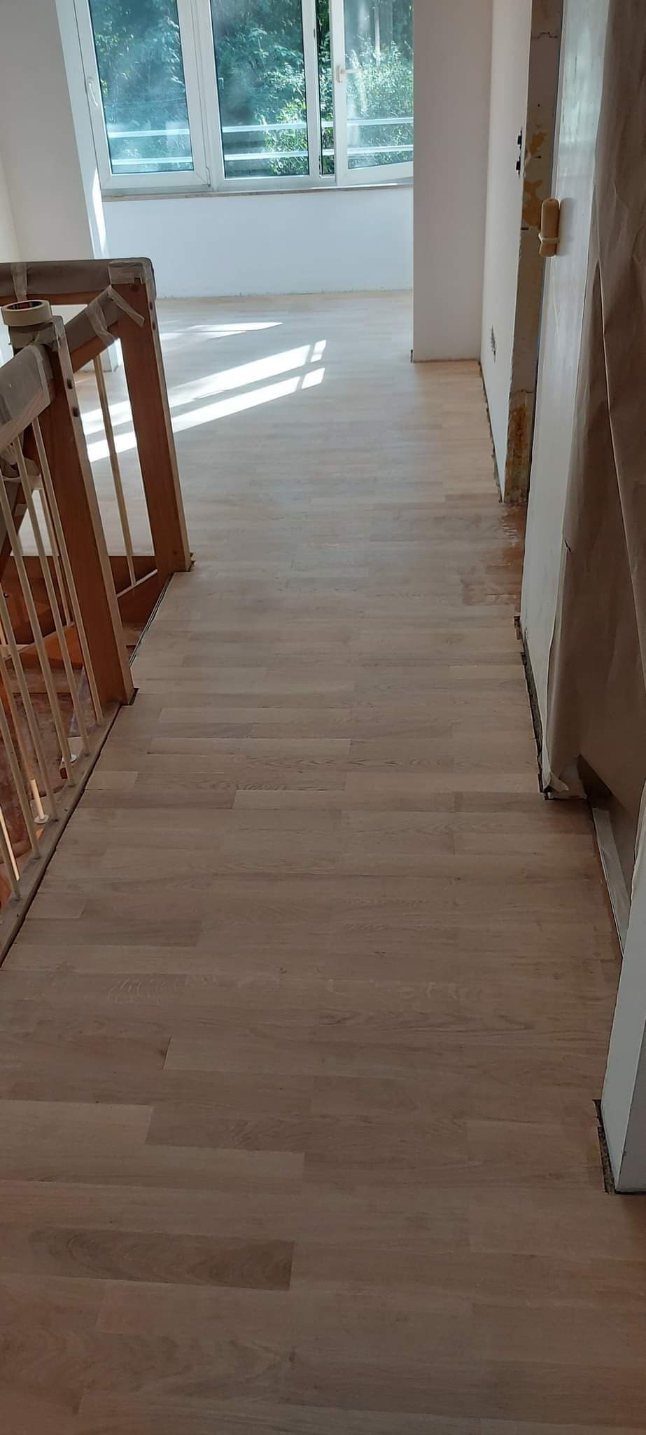 Jasny, nowo ułożony parkiet w korytarzu, widoczne zabezpieczenia na futrynach drzwi i drewnianej balustradzie schodów, wpadające światło dzienne.